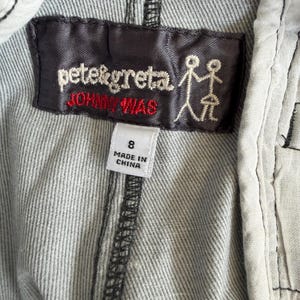 Puede incluir: Una prenda de tela gris con una etiqueta rectangular de color marr&oacute;n oscuro con el texto bordado en blanco pete&greta y el texto en rojo JOHNNY WAS, junto a un icono de figura de palo. Una etiqueta blanca debajo muestra la talla 8 y MADE IN CHINA.