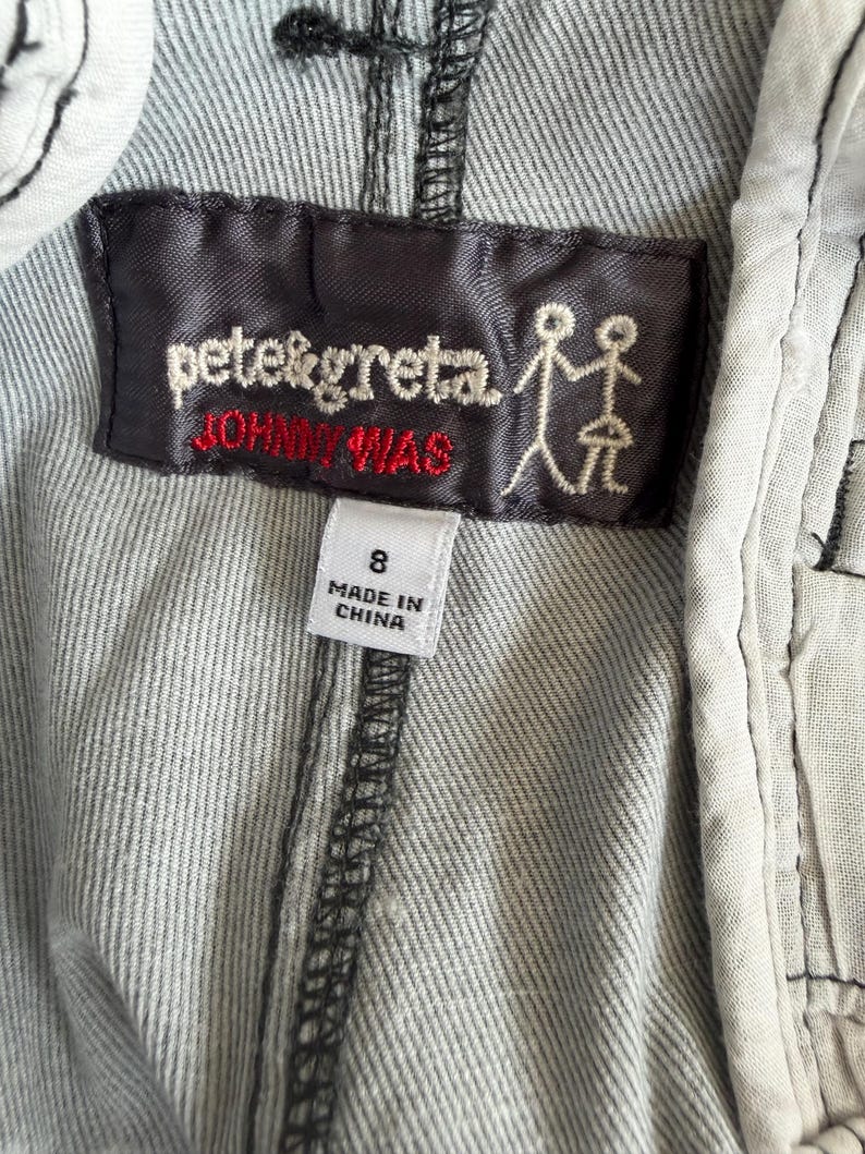 Puede incluir: Una prenda de tela gris con una etiqueta rectangular de color marr&oacute;n oscuro con el texto bordado en blanco pete&greta y el texto en rojo JOHNNY WAS, junto a un icono de figura de palo. Una etiqueta blanca debajo muestra la talla 8 y MADE IN CHINA.