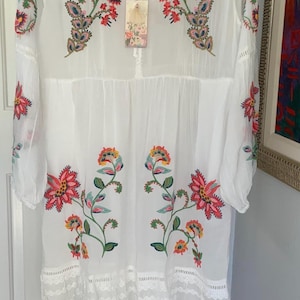 May include: White embroidered dress with colorful floral designs. The dress features long sleeves, a tiered skirt, and a lace hem. A tag with the brand name is attached. The dress is a bohemian style.