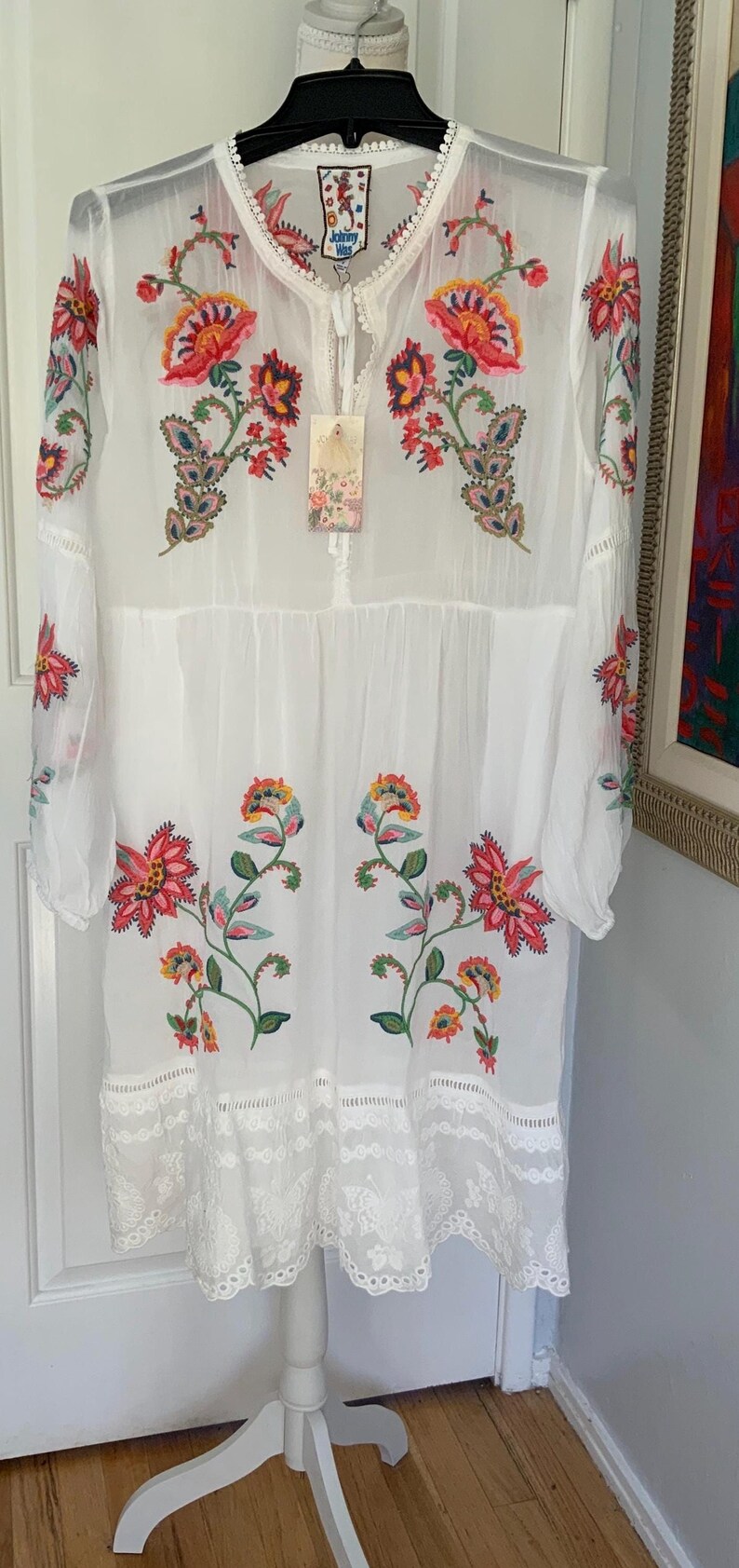 May include: White embroidered dress with colorful floral designs. The dress features long sleeves, a tiered skirt, and a lace hem. A tag with the brand name is attached. The dress is a bohemian style.