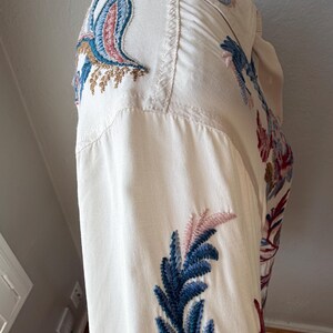 May include: Off-white button-down shirt with intricate embroidery. The embroidery features blue, pink, and brown floral and bird designs. The shirt has a classic collar and buttoned cuffs, showcasing a detailed, artistic style.