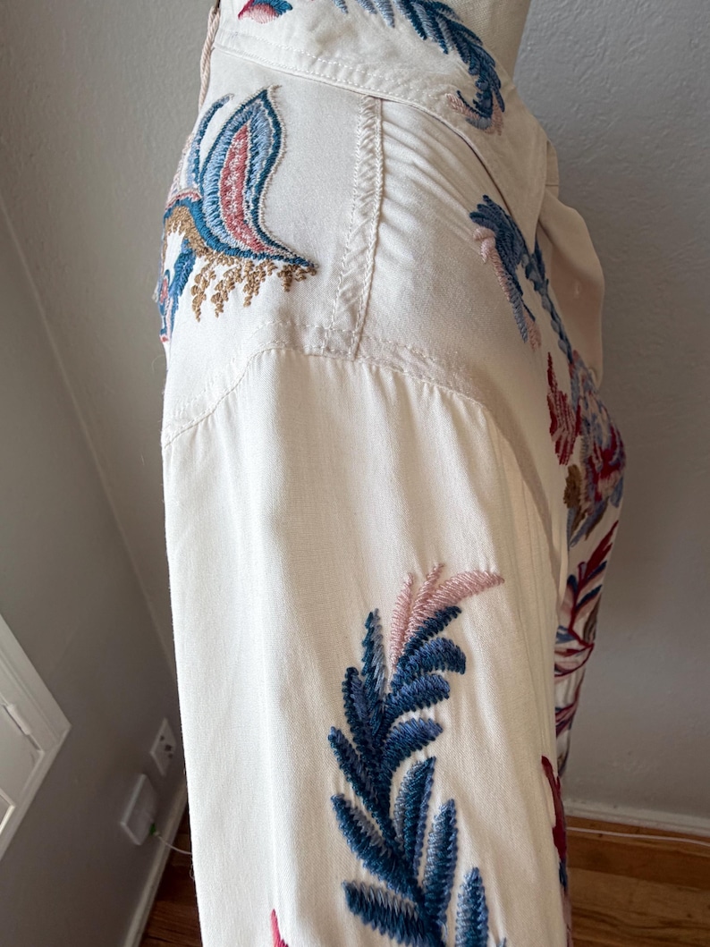 May include: Off-white button-down shirt with intricate embroidery. The embroidery features blue, pink, and brown floral and bird designs. The shirt has a classic collar and buttoned cuffs, showcasing a detailed, artistic style.