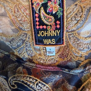 May include: Close-up of a black mesh top with intricate gold embroidery and a colorful paisley design. A rectangular label reads "JOHNNY WAS" with a floral and paisley motif. The label also indicates the size is "S" and "MADE IN CHINA."