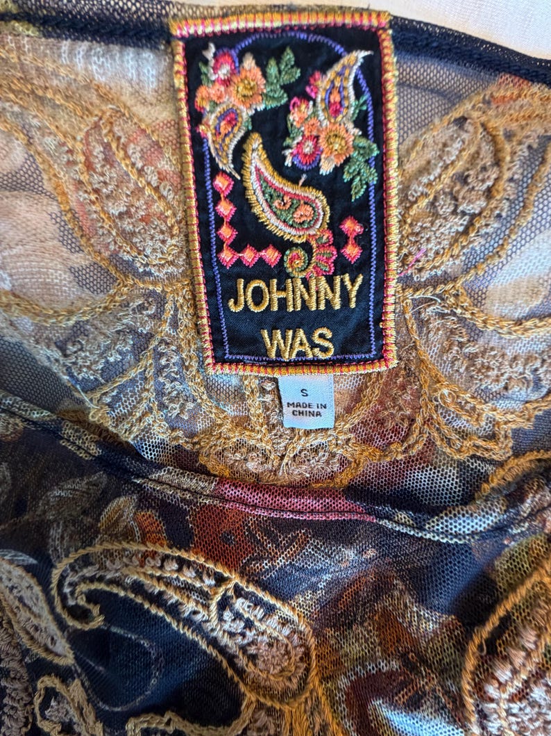 May include: Close-up of a black mesh top with intricate gold embroidery and a colorful paisley design. A rectangular label reads "JOHNNY WAS" with a floral and paisley motif. The label also indicates the size is "S" and "MADE IN CHINA."