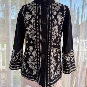 K&ouml;nnte beinhalten: Schwarze Jacke mit wei&szlig;en, gestickten Blumen- und Paisley-Mustern. Die Jacke hat einen hohen Kragen, dreiviertel &Auml;rmel und gro&szlig;e schwarze Kn&ouml;pfe vorne. Die detaillierte Stickerei bedeckt die Vorderseite, die &Auml;rmel und den R&uuml;cken der Jacke.