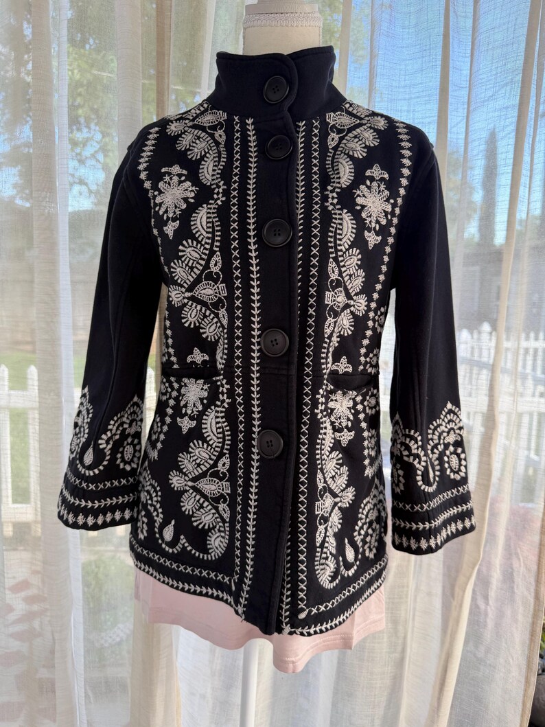 K&ouml;nnte beinhalten: Schwarze Jacke mit wei&szlig;en, gestickten Blumen- und Paisley-Mustern. Die Jacke hat einen hohen Kragen, dreiviertel &Auml;rmel und gro&szlig;e schwarze Kn&ouml;pfe vorne. Die detaillierte Stickerei bedeckt die Vorderseite, die &Auml;rmel und den R&uuml;cken der Jacke.