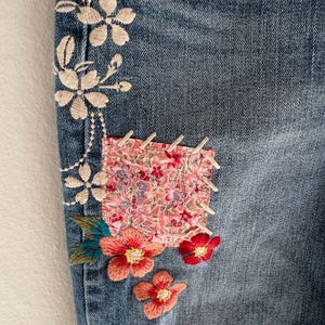 Op de afbeelding: Close-up van blauwe denimstof met bloemenborduurwerk en een stoffen patch. Het borduurwerk heeft witte, koraalrode en rode bloemen met groene bladeren. De patch heeft een roze bloemenpatroon.