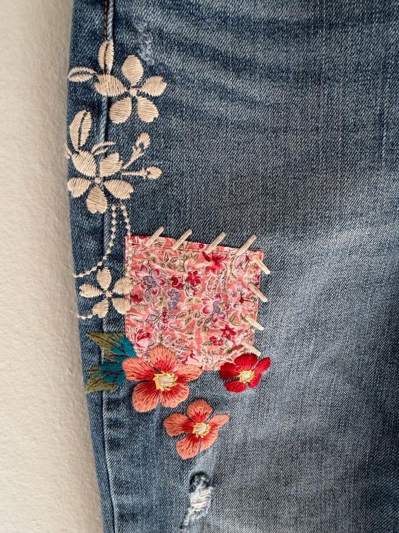 Op de afbeelding: Close-up van blauwe denimstof met bloemenborduurwerk en een stoffen patch. Het borduurwerk heeft witte, koraalrode en rode bloemen met groene bladeren. De patch heeft een roze bloemenpatroon.
