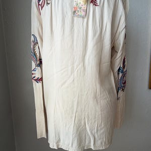 May include: Cream-colored long-sleeve shirt with intricate embroidered paisley and floral designs in shades of blue, red, and green. The shirt has a tag attached. The sleeves have a cuff detail.