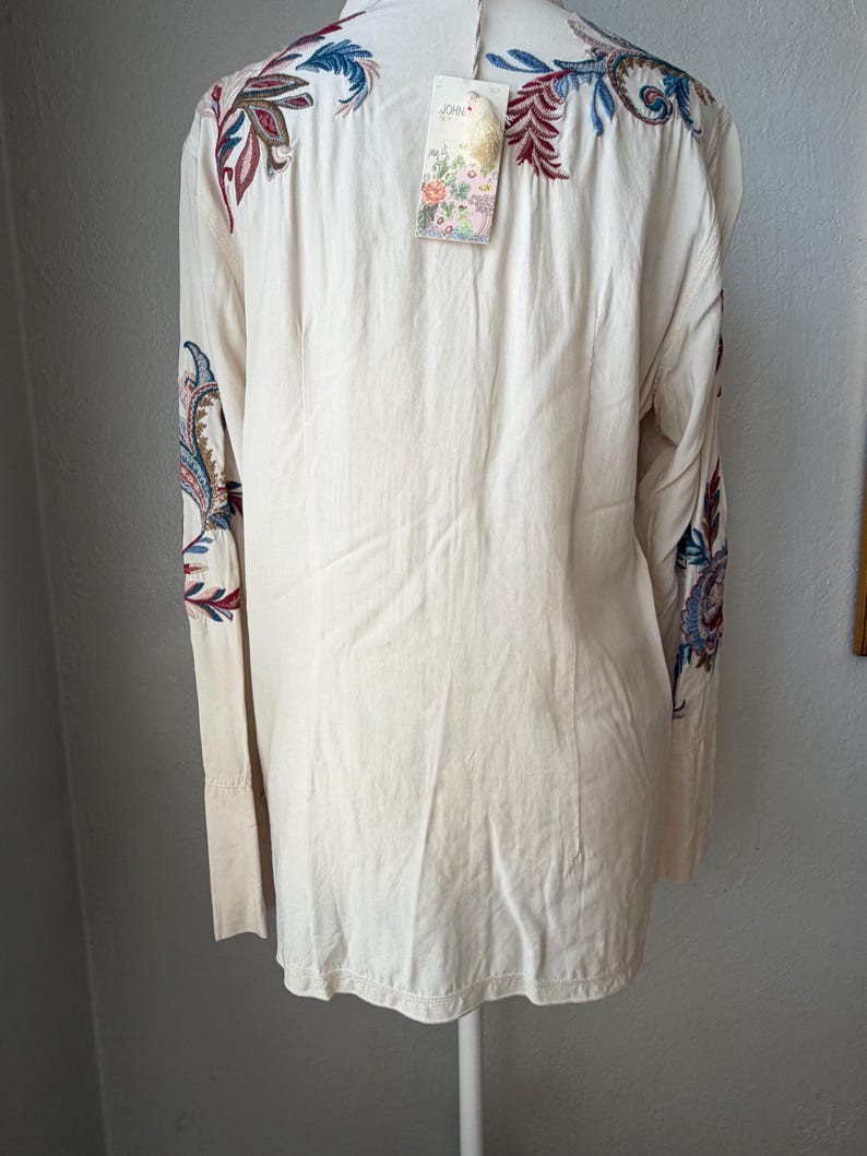 May include: Cream-colored long-sleeve shirt with intricate embroidered paisley and floral designs in shades of blue, red, and green. The shirt has a tag attached. The sleeves have a cuff detail.