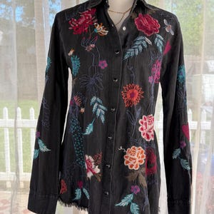 以下が含まれることがあります： カラフルな花の刺繍が施された黒いボタンダウンシャツ。長袖シャツは、赤い、ピンク、オレンジ、ターコイズの花と葉の複雑なデザインと、ほつれた裾が特徴です。「beautiful」という言葉が襟に刺繍されています。