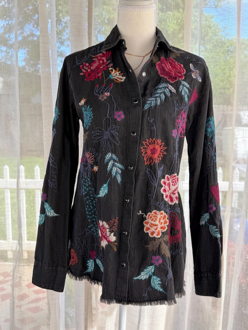 以下が含まれることがあります： カラフルな花の刺繍が施された黒いボタンダウンシャツ。長袖シャツは、赤い、ピンク、オレンジ、ターコイズの花と葉の複雑なデザインと、ほつれた裾が特徴です。「beautiful」という言葉が襟に刺繍されています。