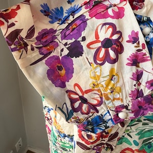 May include: Close-up of a cream-colored garment with a vibrant floral print. The design features large, colorful flowers in shades of purple, pink, red, blue, and yellow. The fabric has pleats and white buttons.
