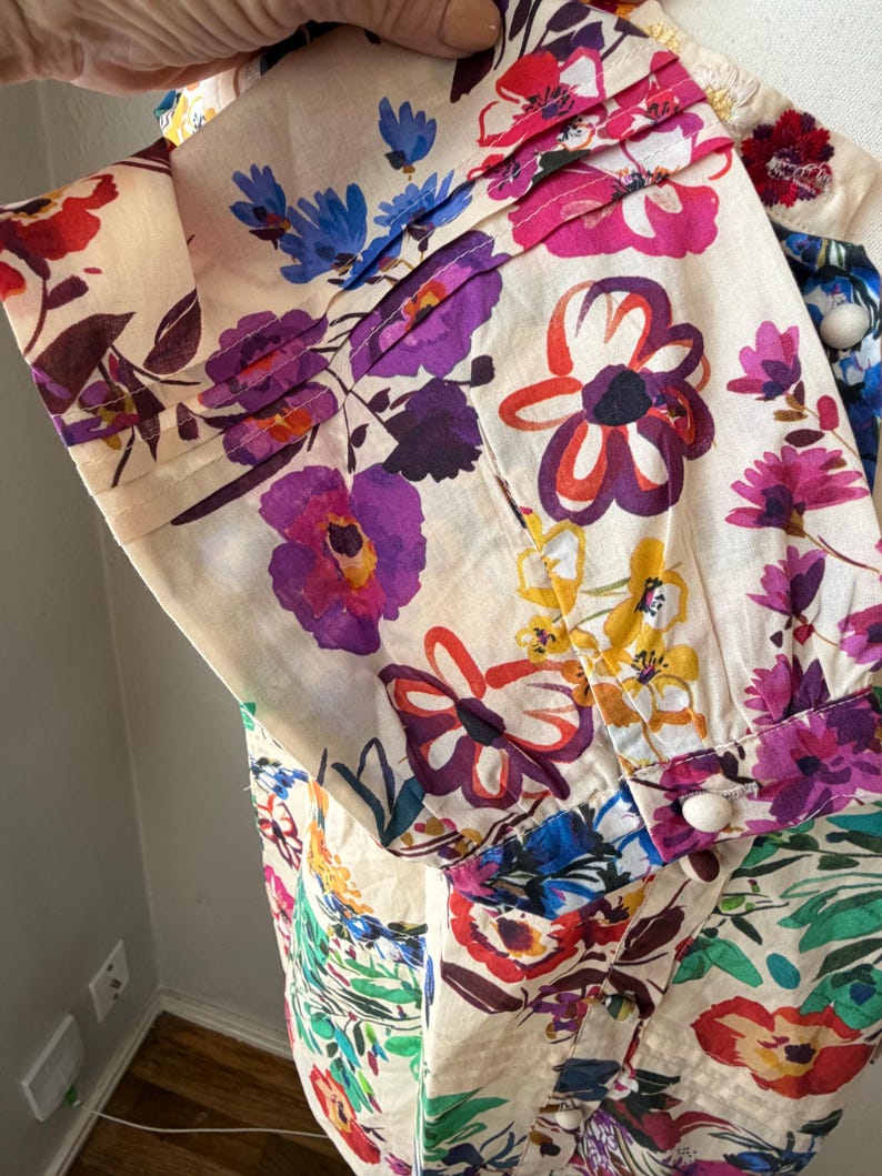 May include: Close-up of a cream-colored garment with a vibrant floral print. The design features large, colorful flowers in shades of purple, pink, red, blue, and yellow. The fabric has pleats and white buttons.
