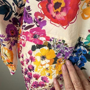 May include: Close-up of a floral print garment featuring large, colorful blooms in shades of red, yellow, purple, and pink. The fabric has a cream-colored background and a patchwork design. The garment appears to be a blouse or shirt.