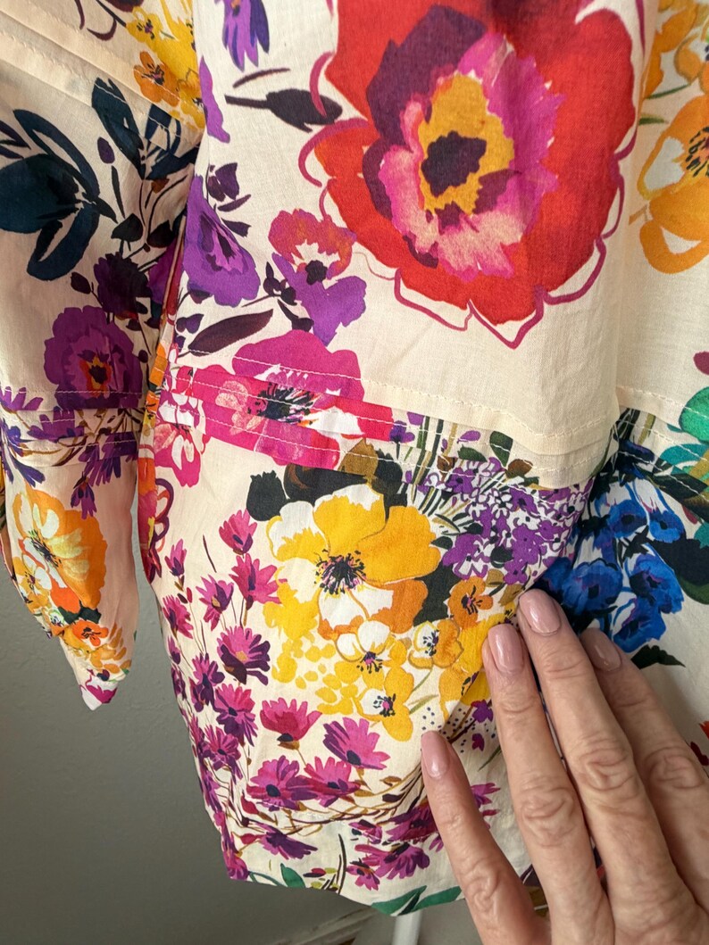 May include: Close-up of a floral print garment featuring large, colorful blooms in shades of red, yellow, purple, and pink. The fabric has a cream-colored background and a patchwork design. The garment appears to be a blouse or shirt.