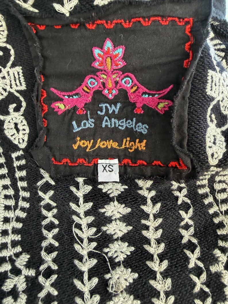 K&ouml;nnte beinhalten: Schwarz besticktes Oberteil mit wei&szlig;en Blumen- und geometrischen Mustern. Ein schwarzes Etikett zeigt farbenfrohe Stickereien von V&ouml;geln und Blumen mit dem Text "JW Los Angeles Joy Love Light". Das Gr&ouml;&szlig;enetikett lautet "XS".