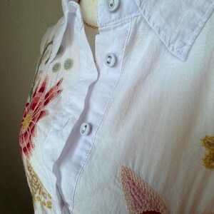 May include: White button-down shirt with colorful floral embroidery. The embroidery features red, pink, yellow, and green thread. The shirt has a collar and three visible buttons. A gold necklace is also visible.