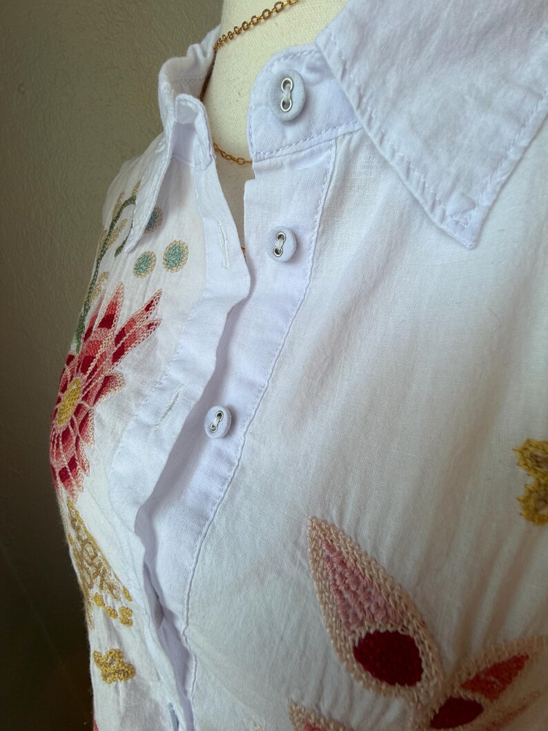 May include: White button-down shirt with colorful floral embroidery. The embroidery features red, pink, yellow, and green thread. The shirt has a collar and three visible buttons. A gold necklace is also visible.