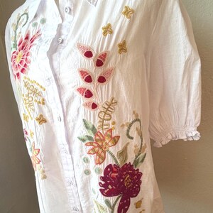 May include: White button-down shirt with short sleeves and floral embroidery. The embroidery features red, pink, yellow, and green flowers and leaves. The shirt has a collared neckline and is displayed on a mannequin.