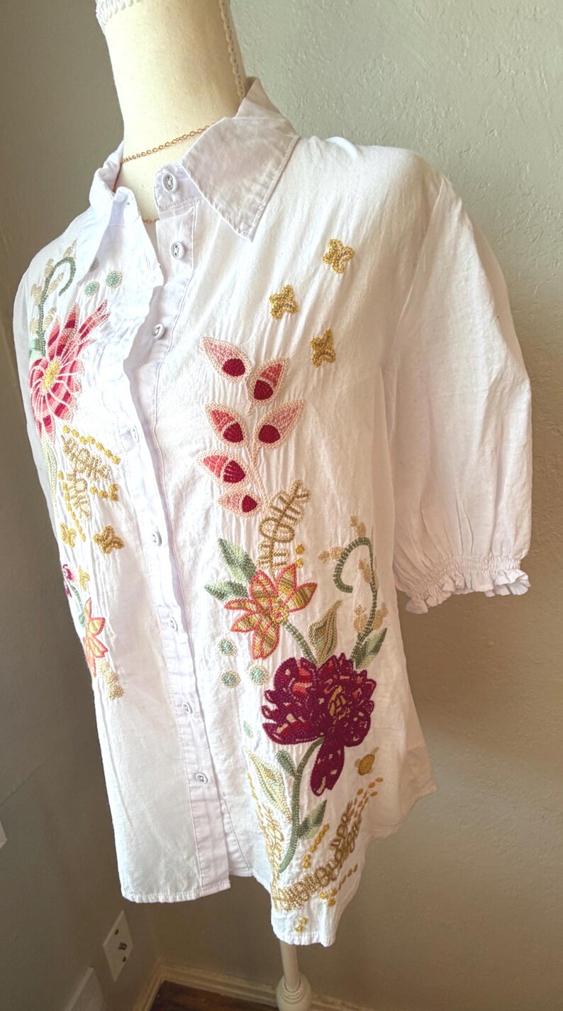 May include: White button-down shirt with short sleeves and floral embroidery. The embroidery features red, pink, yellow, and green flowers and leaves. The shirt has a collared neckline and is displayed on a mannequin.