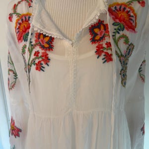 May include: White embroidered tunic with colorful floral designs in red, yellow, and blue. The tunic features a V-neckline with a drawstring and long sleeves. The embroidery is detailed and covers the neckline and sleeves.