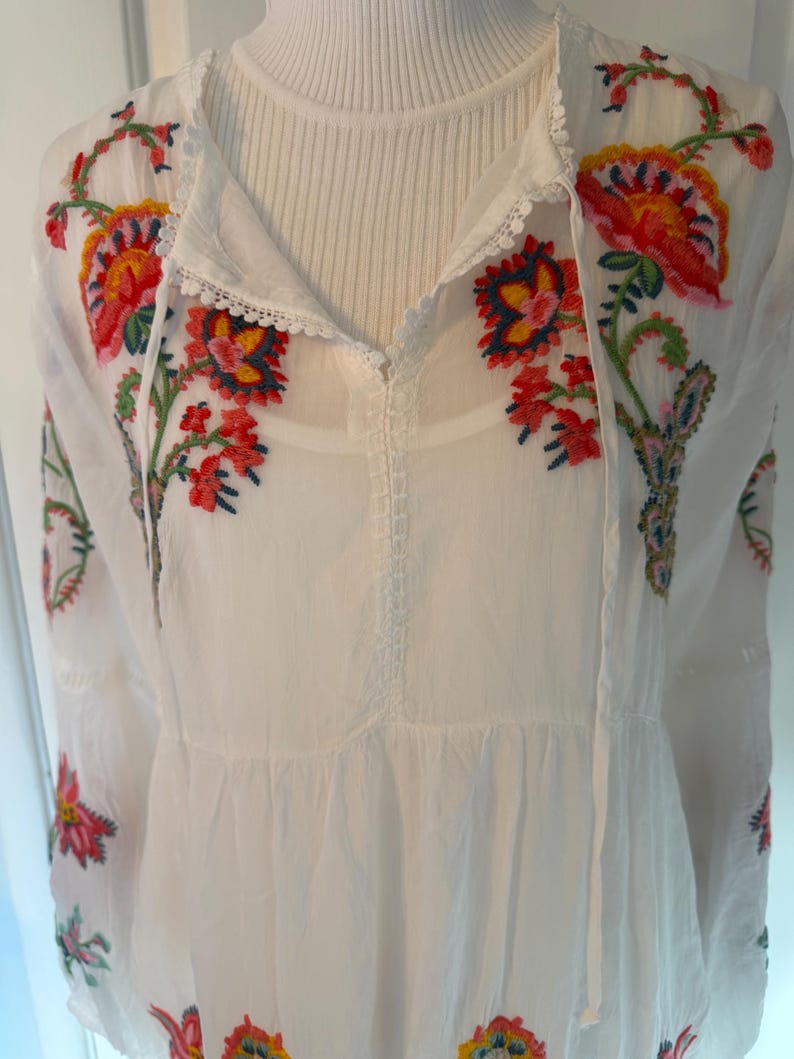 May include: White embroidered tunic with colorful floral designs in red, yellow, and blue. The tunic features a V-neckline with a drawstring and long sleeves. The embroidery is detailed and covers the neckline and sleeves.