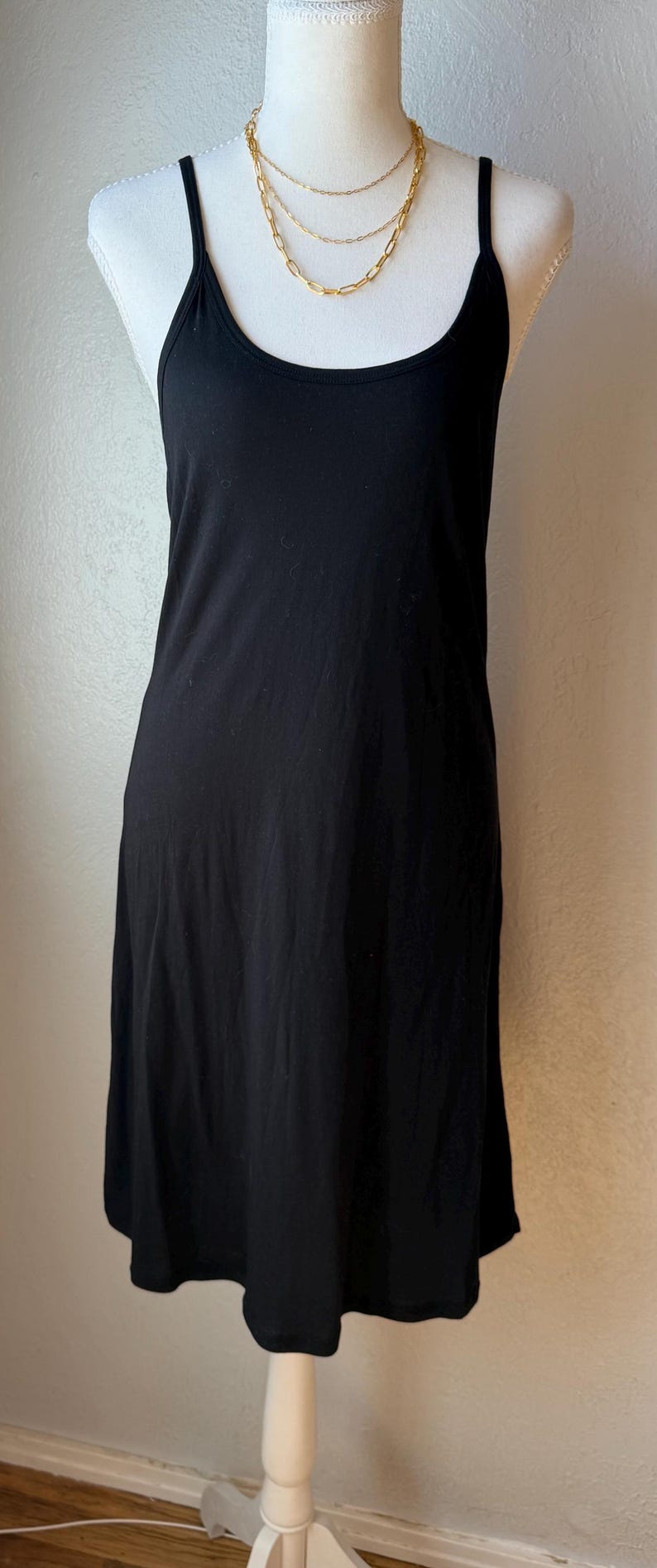 May include: A black slip dress with spaghetti straps is displayed on a white mannequin. A gold-colored layered necklace is draped around the mannequin's neck. The dress has a simple, classic design.