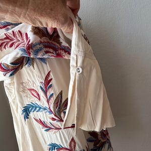 May include: Cream-colored shirt with floral embroidery in shades of red, blue, and brown. The embroidery is detailed and covers the front of the shirt. The sleeve is rolled up and buttoned.