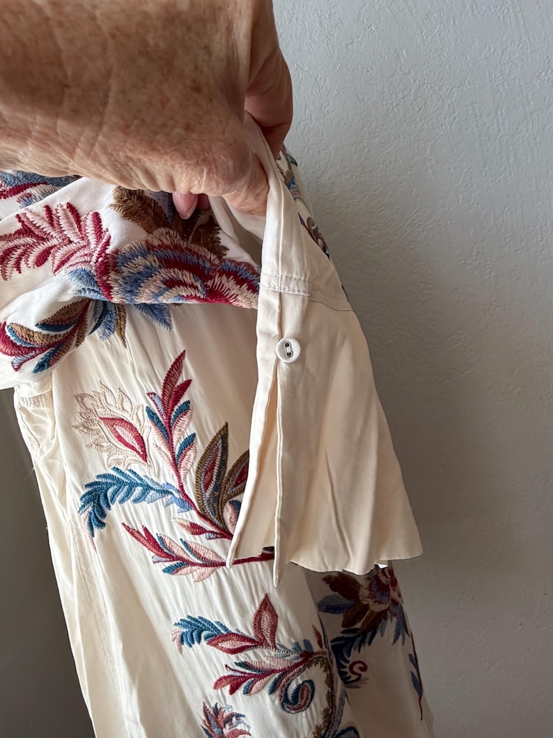 May include: Cream-colored shirt with floral embroidery in shades of red, blue, and brown. The embroidery is detailed and covers the front of the shirt. The sleeve is rolled up and buttoned.