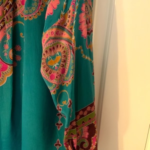 May include: A teal-colored long-sleeved top with a vibrant paisley pattern in pink, orange, and brown. The intricate design features floral and heart-shaped motifs, creating a bohemian aesthetic. The sleeve cuffs have a darker brown border with a floral design.