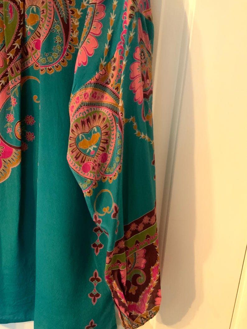 May include: A teal-colored long-sleeved top with a vibrant paisley pattern in pink, orange, and brown. The intricate design features floral and heart-shaped motifs, creating a bohemian aesthetic. The sleeve cuffs have a darker brown border with a floral design.