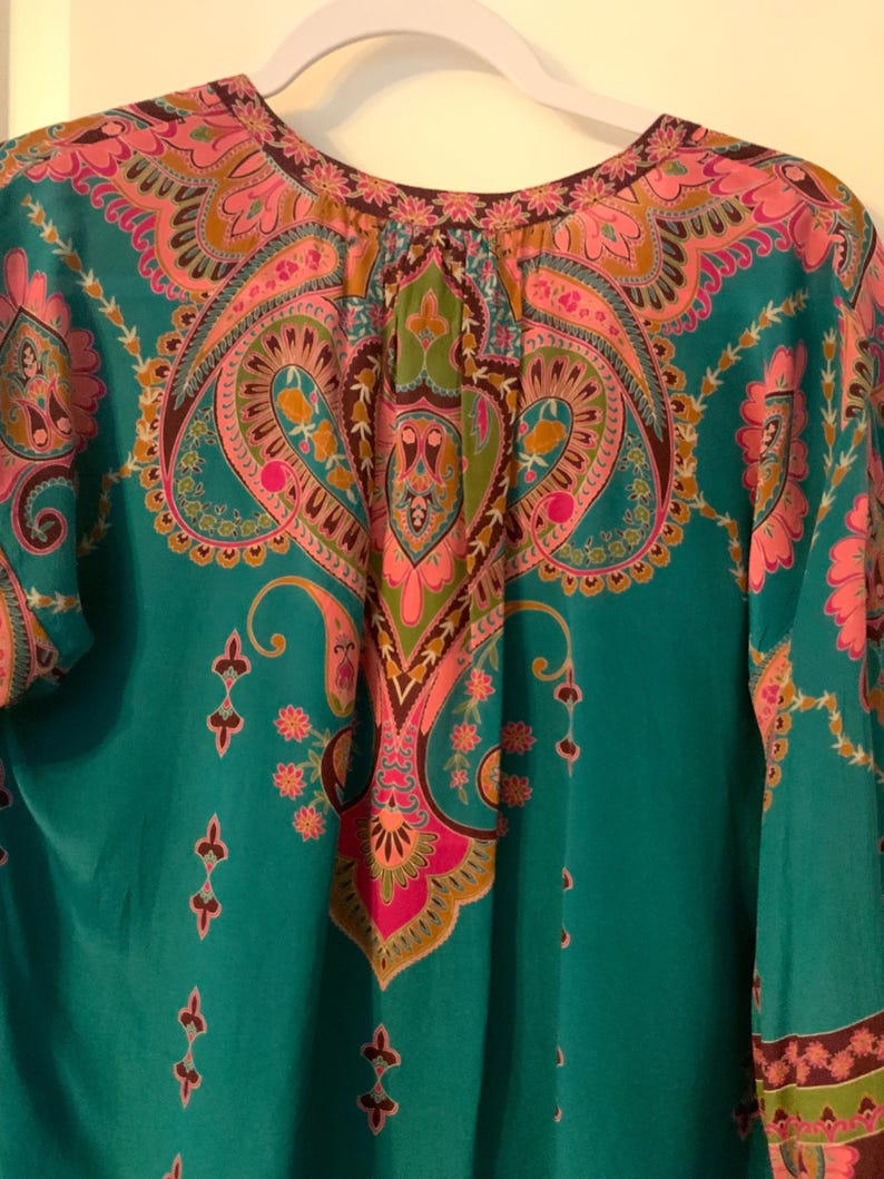 May include: A teal-colored top with a vibrant paisley pattern in pink, orange, and green. The top features a round neckline and long sleeves. The intricate design includes floral and teardrop motifs.