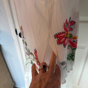 May include: White dress with colorful floral embroidery. The embroidery features red, pink, and green flowers with blue accents. The dress has a scalloped hem and eyelet detailing.