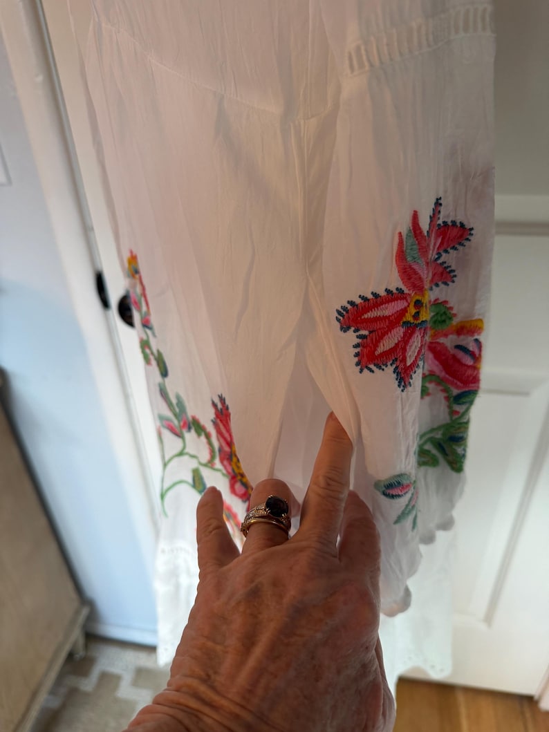 May include: White dress with colorful floral embroidery. The embroidery features red, pink, and green flowers with blue accents. The dress has a scalloped hem and eyelet detailing.