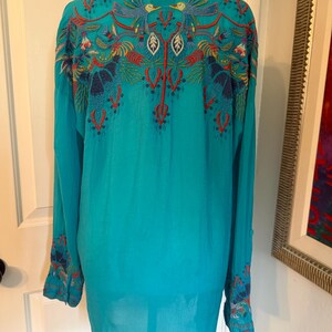 May include: A turquoise long-sleeve shirt with intricate embroidery on the back and cuffs. The embroidery features birds, leaves, and floral patterns in red, blue, and yellow. The shirt has a button-down collar.