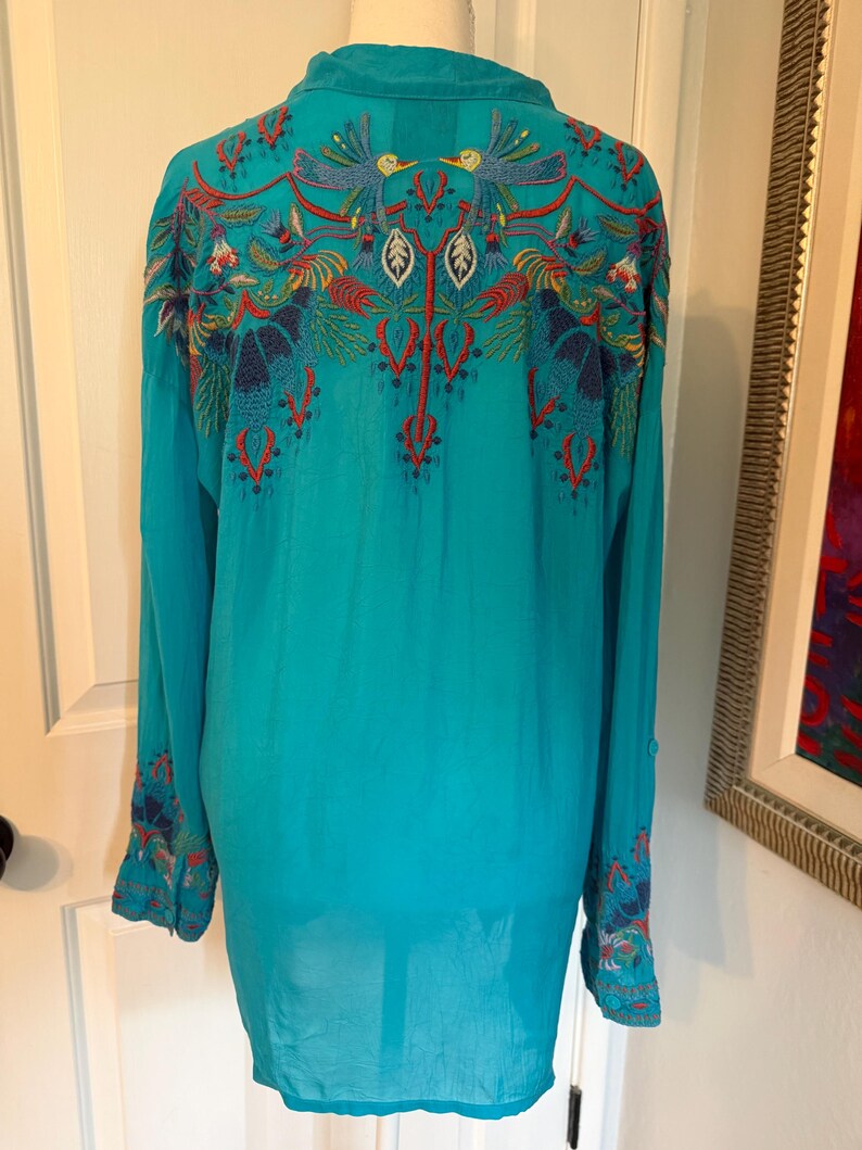 May include: A turquoise long-sleeve shirt with intricate embroidery on the back and cuffs. The embroidery features birds, leaves, and floral patterns in red, blue, and yellow. The shirt has a button-down collar.