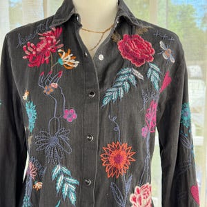 以下が含まれることがあります： カラフルな花の刺繍が施された、濃いグレーのボタンダウンシャツ。 刺繍にはピンク、赤、オレンジ、ターコイズの花と葉が使われています。 シャツは長袖で、襟が付いています。 ゴールドのネックレスが見えます。
