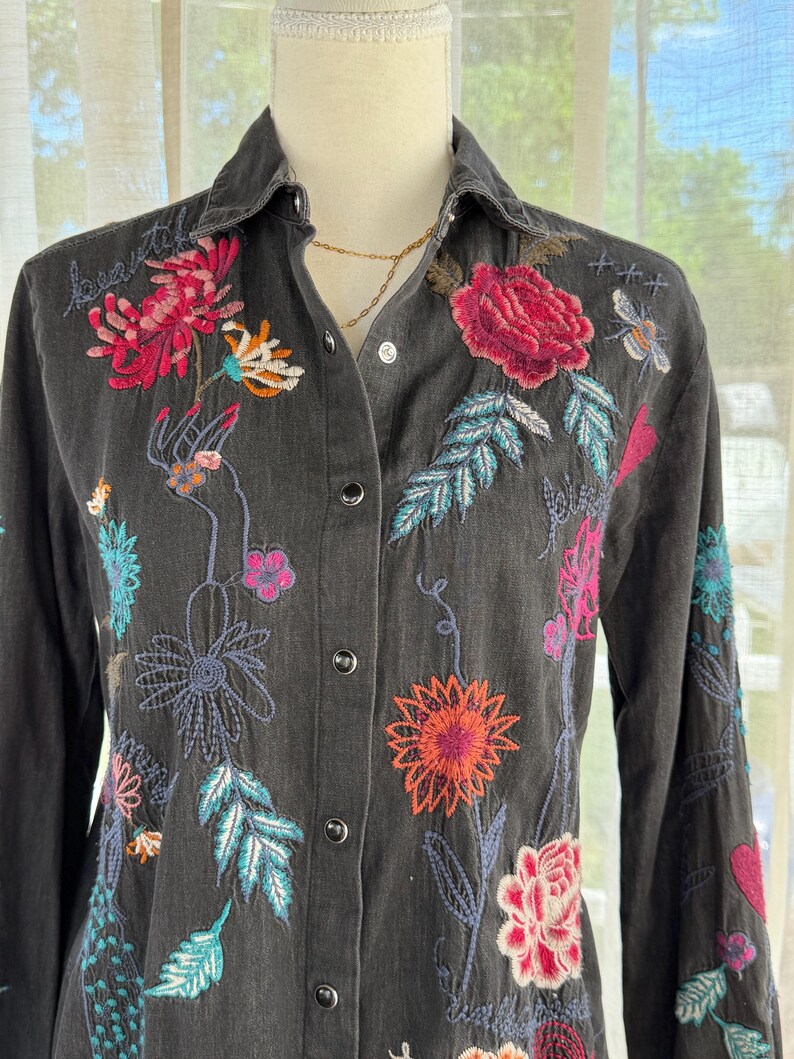 以下が含まれることがあります： カラフルな花の刺繍が施された、濃いグレーのボタンダウンシャツ。 刺繍にはピンク、赤、オレンジ、ターコイズの花と葉が使われています。 シャツは長袖で、襟が付いています。 ゴールドのネックレスが見えます。