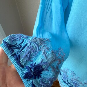 Puede incluir: Una blusa azul claro con detalles bordados. El pu&ntilde;o de la manga presenta intrincados patrones florales y de hojas en tonos azules. La tela parece ligera y fluida, con una textura ligeramente arrugada. La blusa tiene un corte relajado.
