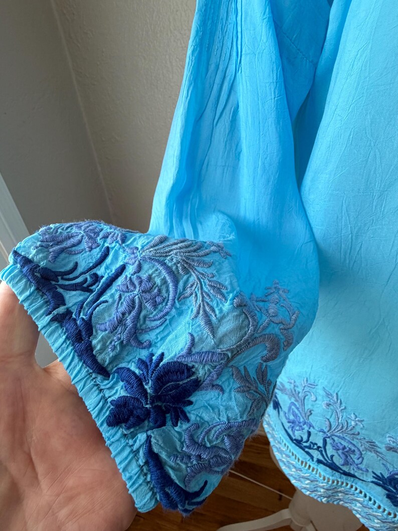 Puede incluir: Una blusa azul claro con detalles bordados. El pu&ntilde;o de la manga presenta intrincados patrones florales y de hojas en tonos azules. La tela parece ligera y fluida, con una textura ligeramente arrugada. La blusa tiene un corte relajado.