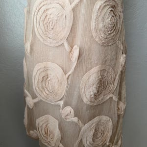 May include: A light beige dress with a textured surface and large, circular floral appliqu&eacute;s. The appliqu&eacute;s are a lighter shade of beige and are arranged in a repeating pattern across the dress.