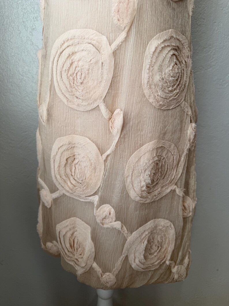 May include: A light beige dress with a textured surface and large, circular floral appliqu&eacute;s. The appliqu&eacute;s are a lighter shade of beige and are arranged in a repeating pattern across the dress.