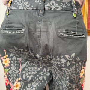 Puede incluir: Pantalones negros con un patr&oacute;n floral y de cachemira en gris oscuro. Los pantalones incluyen una cintura con trabillas, dos bolsillos traseros ribeteados y un estampado floral vibrante en tonos rosa, naranja y amarillo en las costuras laterales.