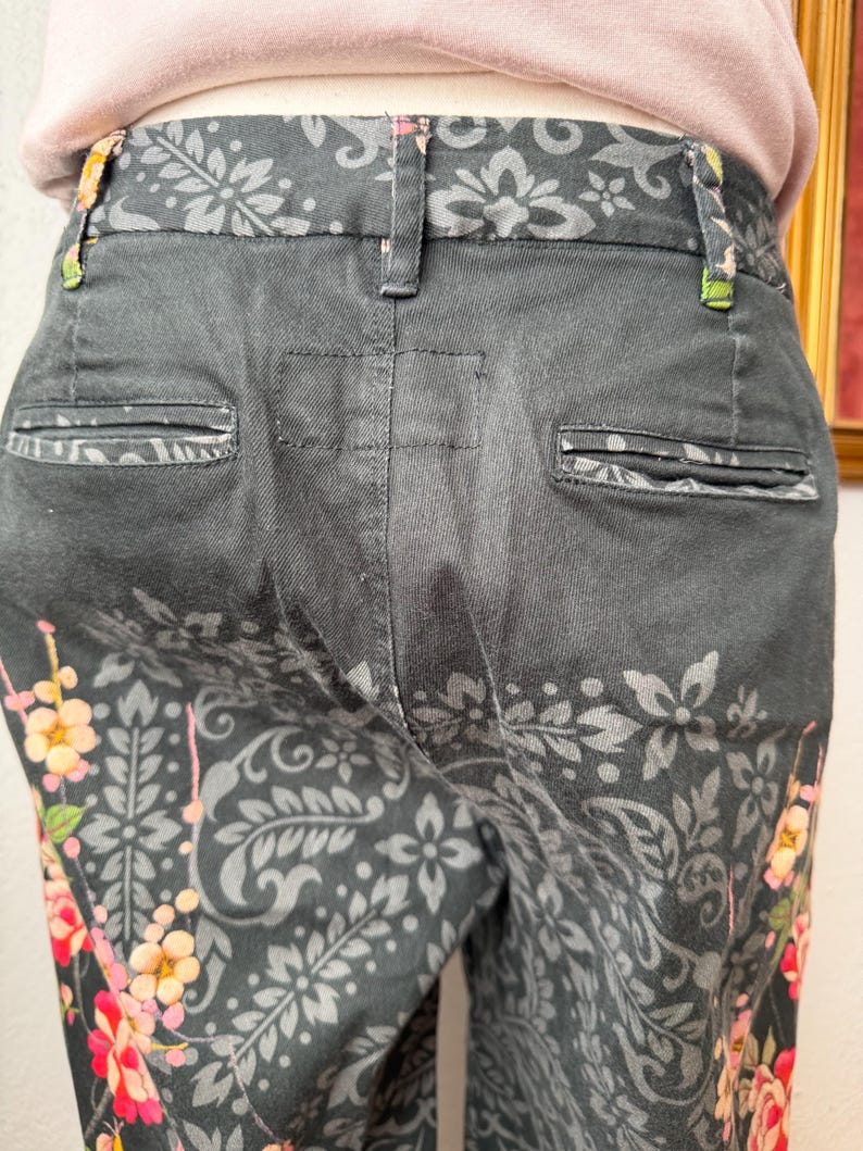 Puede incluir: Pantalones negros con un patr&oacute;n floral y de cachemira en gris oscuro. Los pantalones incluyen una cintura con trabillas, dos bolsillos traseros ribeteados y un estampado floral vibrante en tonos rosa, naranja y amarillo en las costuras laterales.