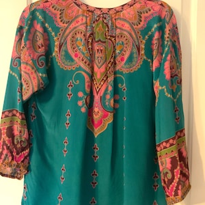 May include: Teal-colored blouse with a vibrant paisley pattern in pink, orange, and green. The long-sleeved top features a round neckline and decorative details. The sleeves have a patterned cuff.