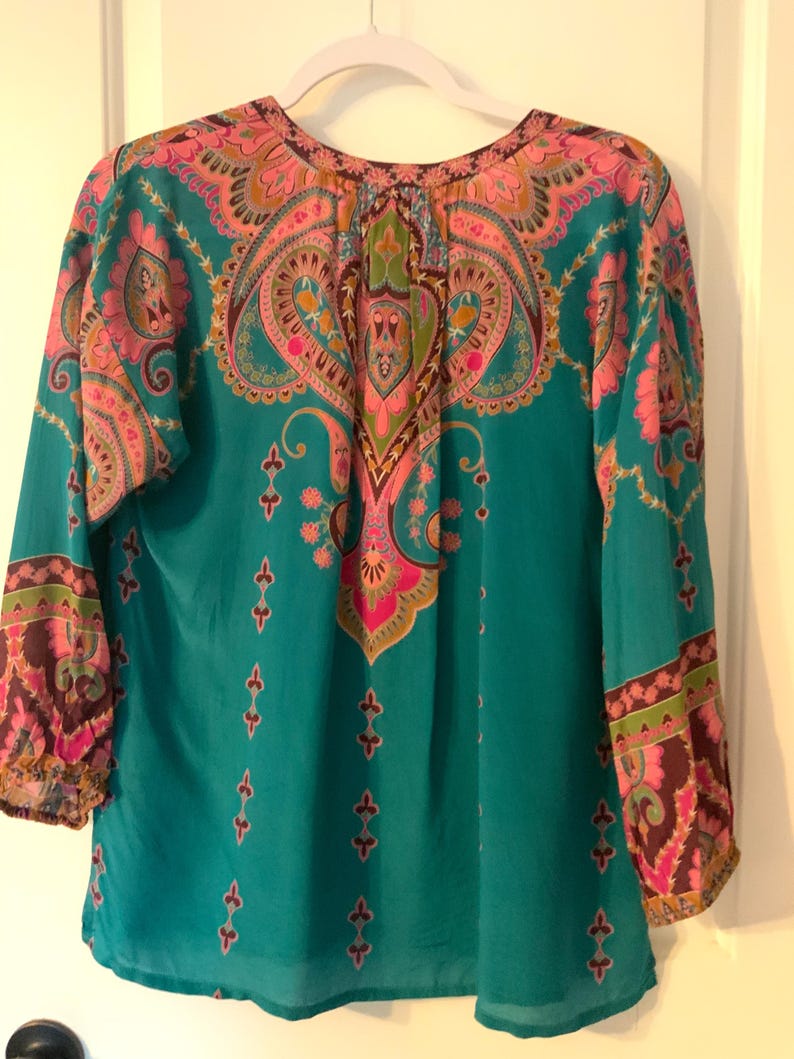 May include: Teal-colored blouse with a vibrant paisley pattern in pink, orange, and green. The long-sleeved top features a round neckline and decorative details. The sleeves have a patterned cuff.