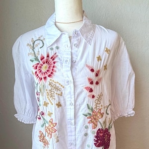 May include: White button-down shirt with short sleeves and floral embroidery in shades of red, pink, and gold. The embroidered flowers and leaves are scattered across the front of the shirt.