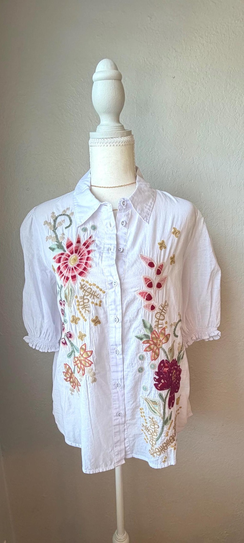 May include: White button-down shirt with short sleeves and floral embroidery in shades of red, pink, and gold. The embroidered flowers and leaves are scattered across the front of the shirt.