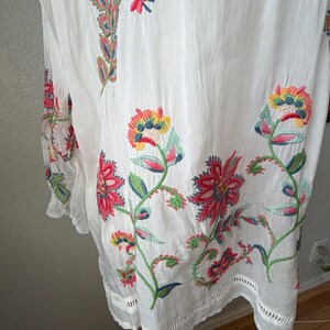 May include: White tunic with colorful floral embroidery. The embroidery features red, pink, yellow, and green flowers and leaves. The tunic has a loose fit and a scalloped hemline. The fabric appears to be lightweight and slightly sheer.